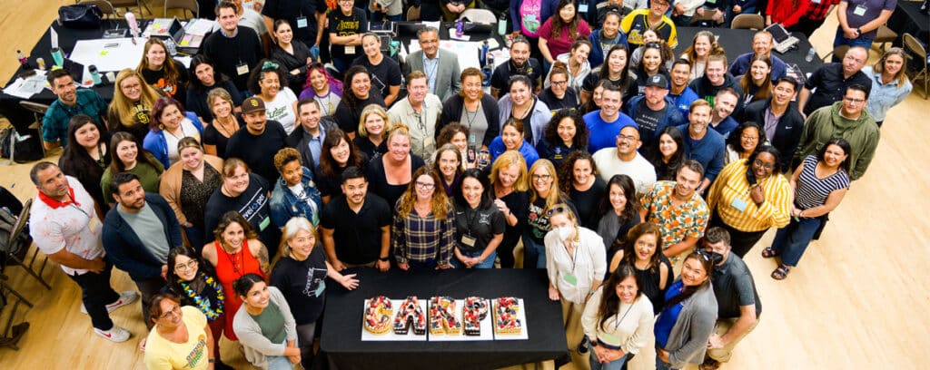 A large, diverse group of people stand closely together in a bright room, smiling up at the camera. In front of them is a table with a cake that spells out UNITY in colorful letters.