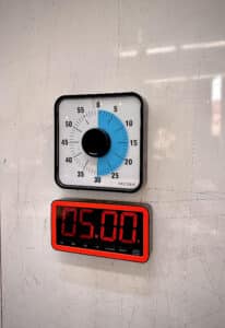 A mechanical timer set to 25 minutes is mounted above a digital clock displaying 05:00, both attached to a whiteboard