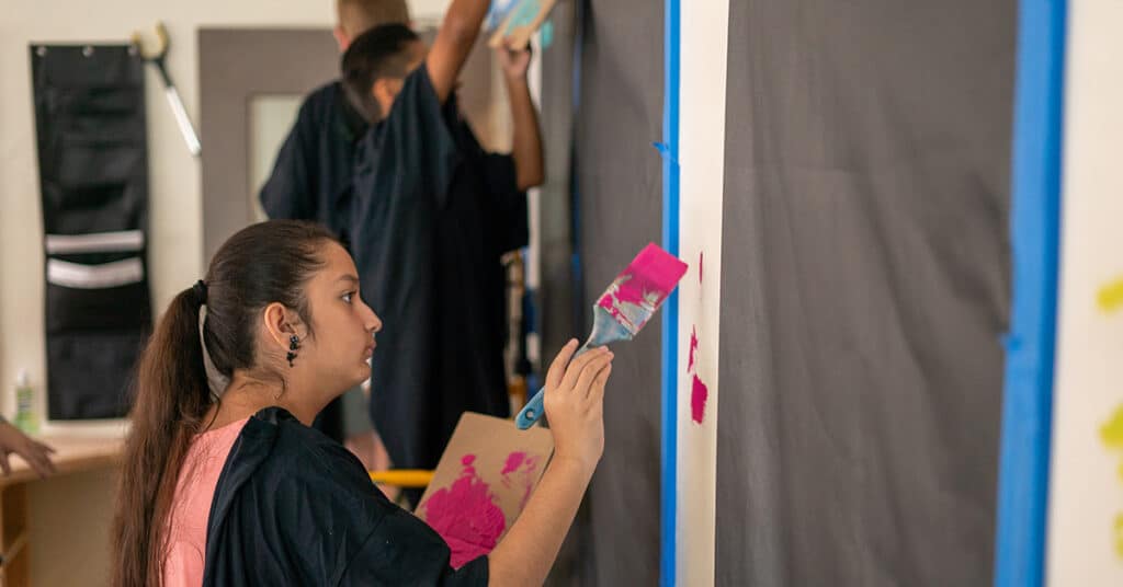 A young person holds a paintbrush with bright pink paint, painting on a wall covered in black paper, while others collaborate in the background, highlighting the spirit of group work during the creative process.