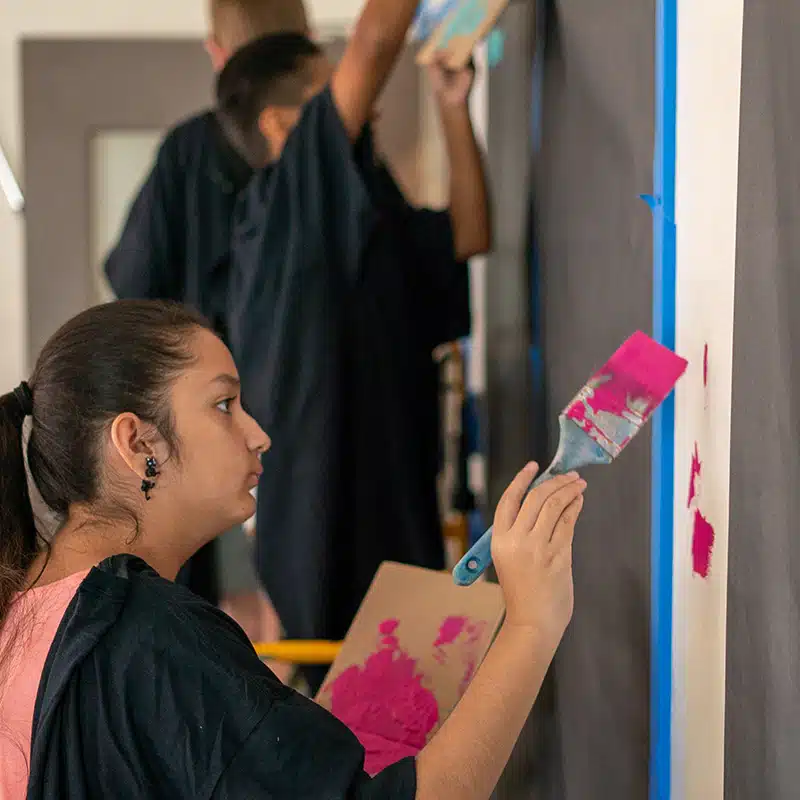 A young person holds a paintbrush and paints magenta patches on a white wall, while others in the background join in. All wear black aprons or shirts to protect their clothing, showcasing collaboration through creative group work.