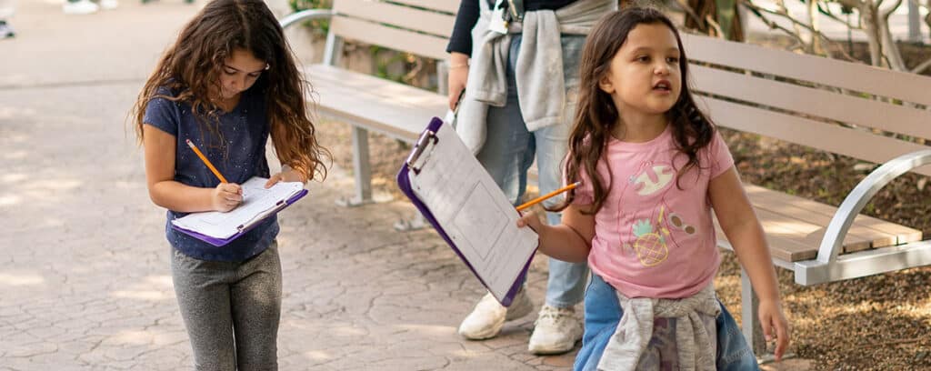 Two young girls holding clipboards and pencils walk through an outdoor area, engaged in a learning activity.