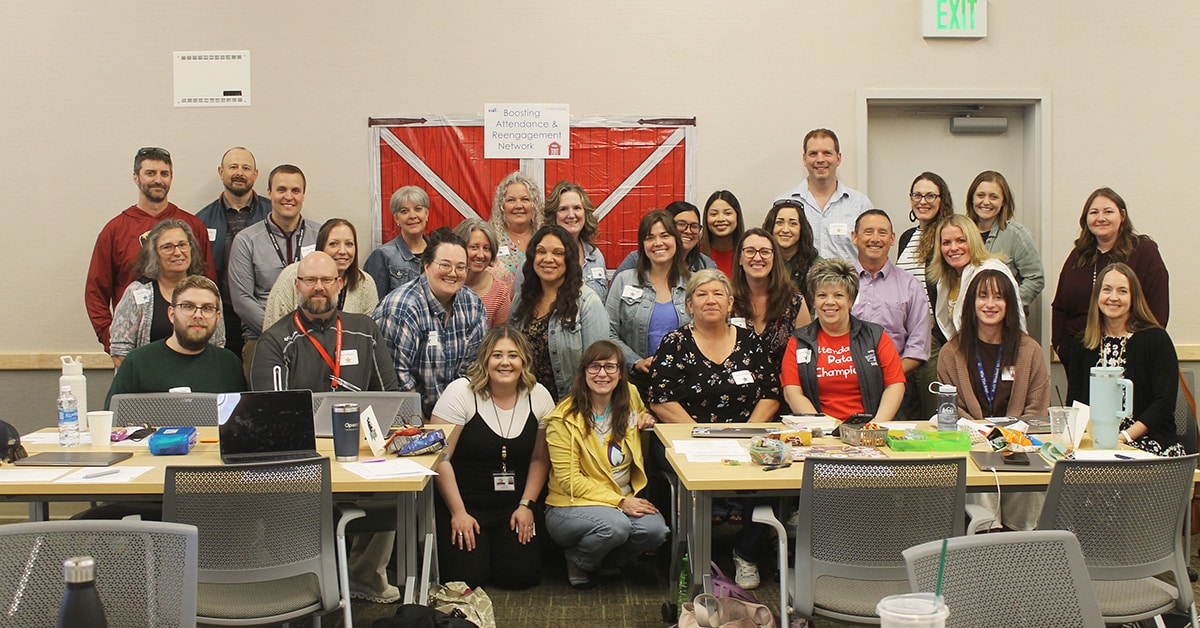 A large group of adults pose together in a classroom setting, some sitting at tables with laptops and snacks, others standing behind, smiling at the camera—a gathering focused on school improvement networks. A red and white banner is displayed on the back wall.