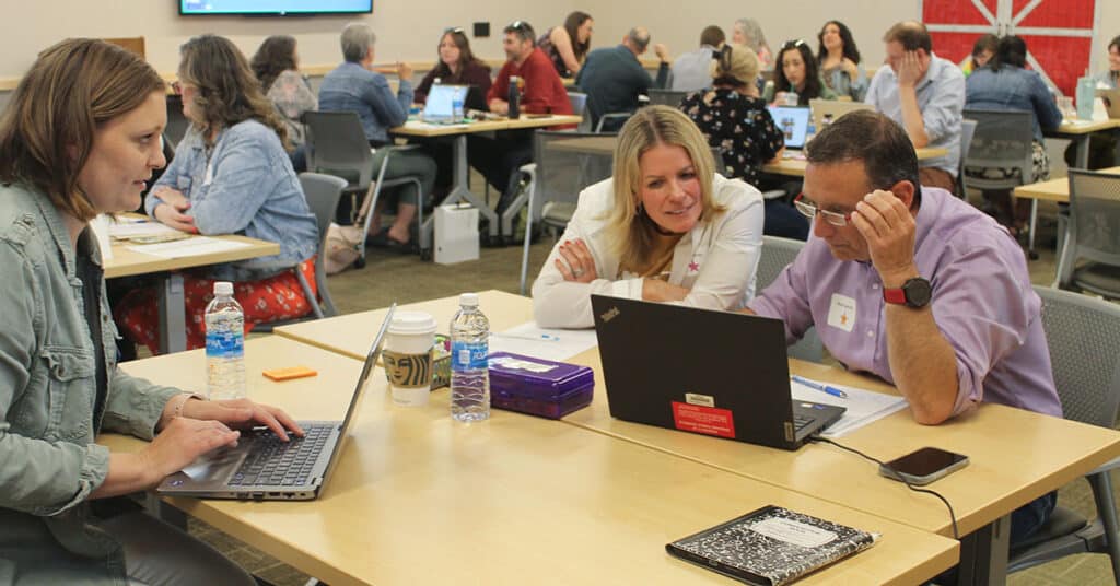 Several people sit at tables in a classroom, working together on laptops. Three adults in the foreground share lessons learned, surrounded by notebooks and coffee cups, while more groups collaborate on school improvement projects in the background.
