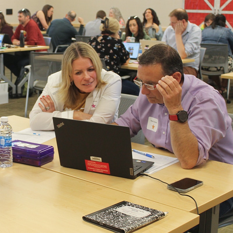 Two people sit at a table collaborating and looking at a laptop screen, engaging in education lessons. Other groups work together in the background, reflecting a dynamic classroom or workshop focused on school improvement.