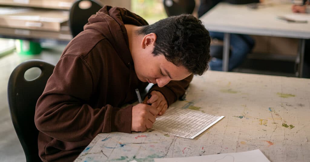 A student wearing a brown hoodie sits at a paint-splattered desk, focusing intently while filling out a quiz with a pen in a classroom setting, preparing for grading.