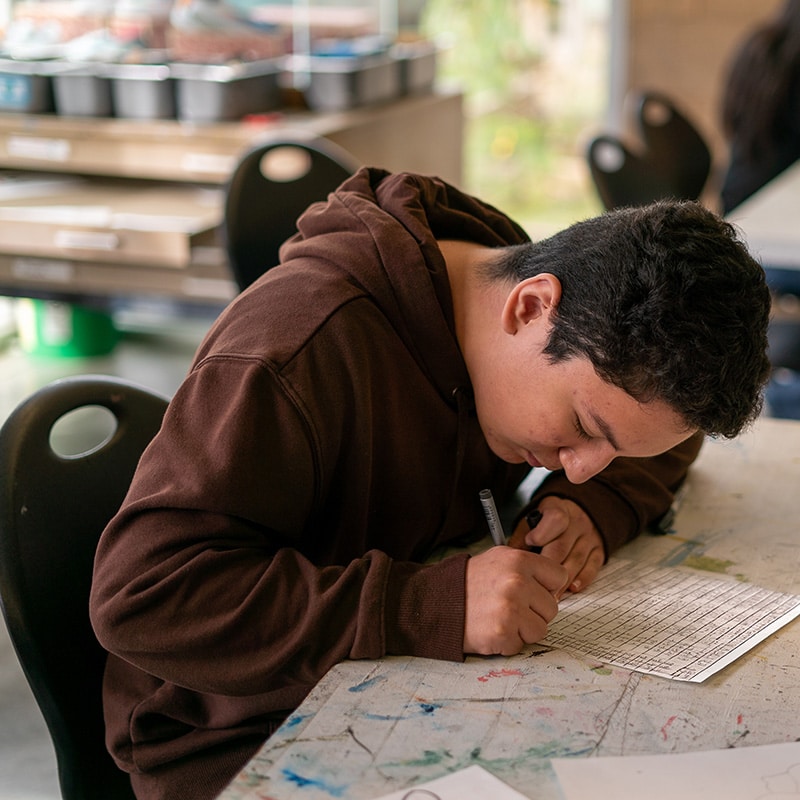 A student in a brown hoodie sits at a table, leaning over and writing on a worksheet in a classroom setting, possibly finishing up quizzes. The table is covered with scattered papers and has a few scribbles or marks on its surface.