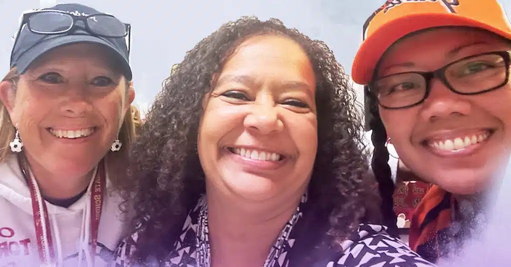 Three smiling women pose for a selfie, capturing a cheerful Bernal episode. The woman in the center has curly hair, while the others wear navy and orange caps, with one sporting glasses—all radiating happiness.