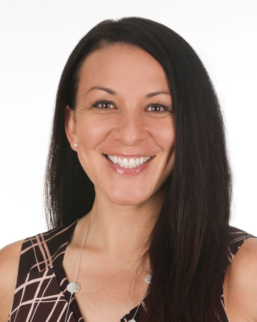 Natalie Daniels, with long dark hair, smiles at the camera. She is wearing a sleeveless patterned top and a silver necklace, set against a white background.