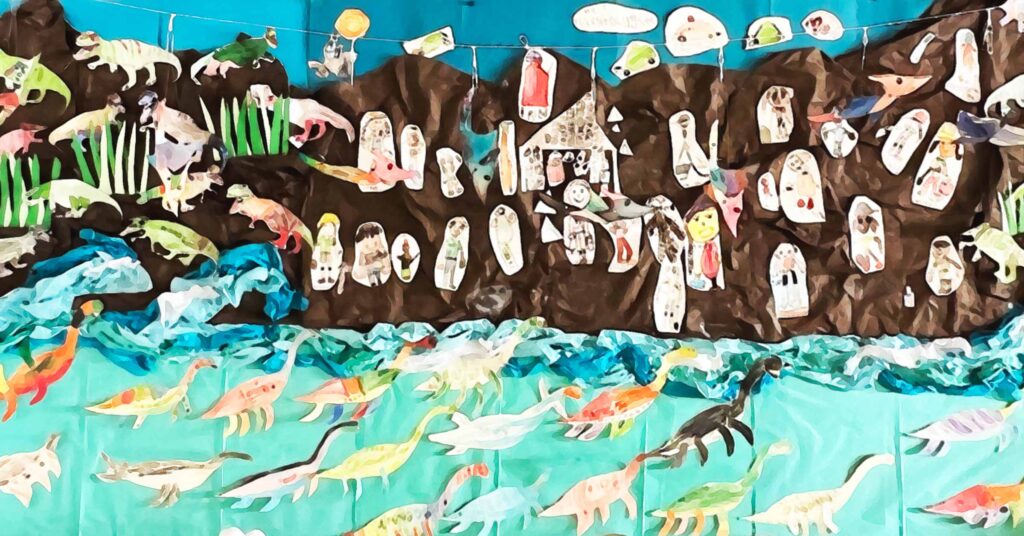 A colorful mural of hand-drawn dinosaurs and people, with dinosaurs in blue water at the bottom and people depicted in front of rocky brown hills and green plants above. The artwork appears to be made by children.