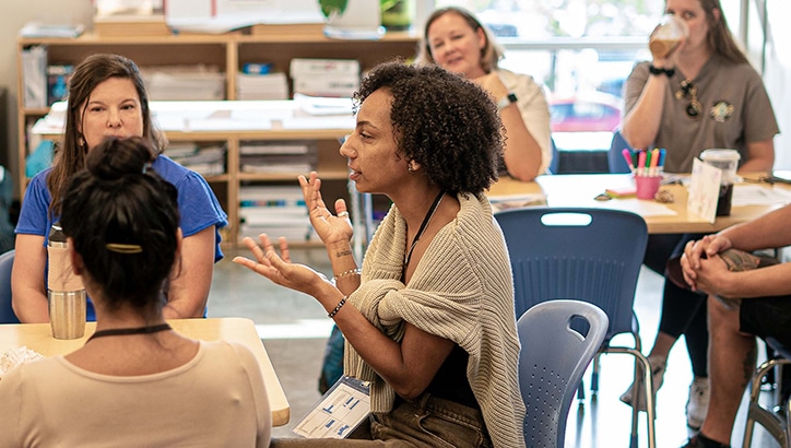 Several adults sit in a classroom, engaged in a group discussion. One woman in the center gestures with her hands while speaking, and others listen attentively. Tables have cups, pens, and papers on them.