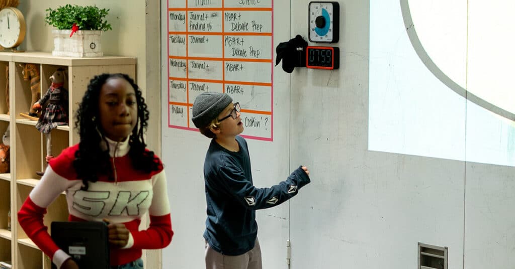 A boy in glasses and a beanie presses a button on a wall timer in the classroom while a girl in a red and white sweater walks by holding a tablet. A whiteboard with schedules and clocks is visible behind them.