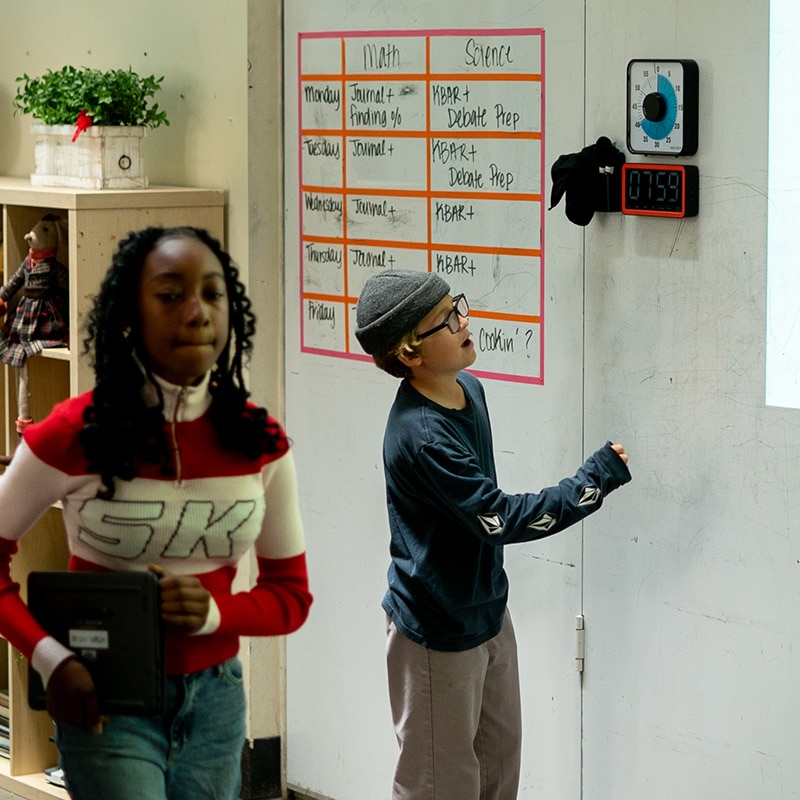 Two children are in a classroom; one girl is walking while holding a tablet, and a boy uses educational tools by pressing a timer on the wall near a schedule chart listing math and science activities.