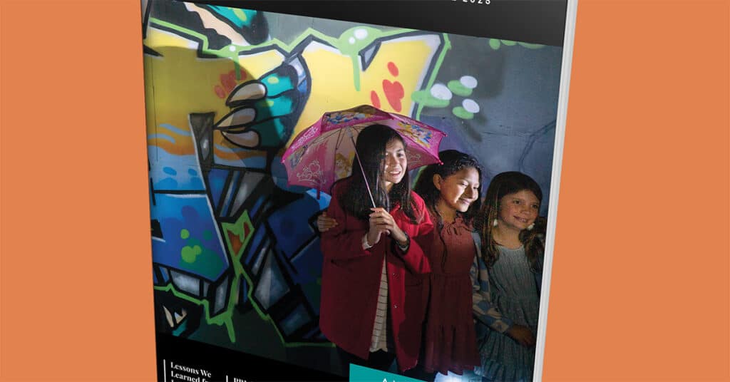 Three girls stand smiling in front of a colorful graffiti wall. The center girl holds a patterned pink umbrella as the lighting highlights their faces, giving Issue 29 Fall 2025 a cheerful, vibrant atmosphere.
