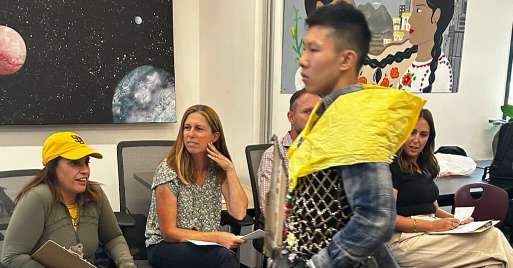 A young person in a yellow and silver costume walks past seated adults, some holding clipboards, in a classroom decorated with colorful murals and space-themed art—against all expectations, it’s clear they truly love school.