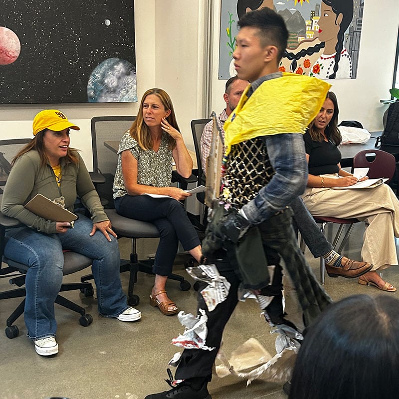 A student models a creative outfit made of recycled materials during a classroom fashion show, while teachers and students—who began to love school—watch and take notes. Colorful artwork hangs on the wall in the background.