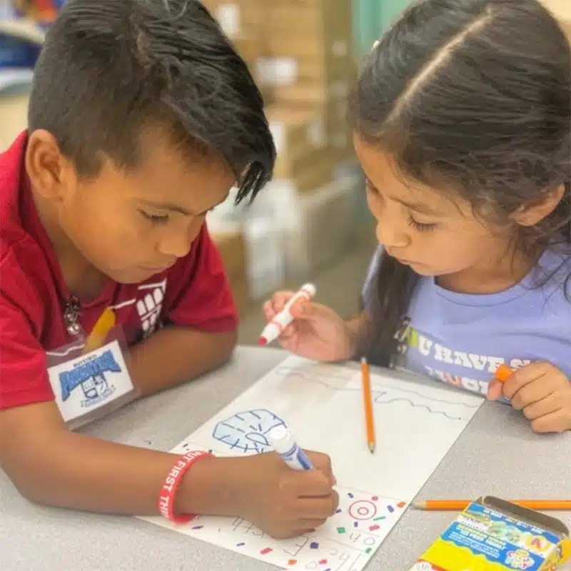 Two young children sit at a table, drawing and coloring on paper with markers and pencils. They appear focused and engaged in their creative activity—a joyful step toward literacy—while a box of crayons is visible nearby.