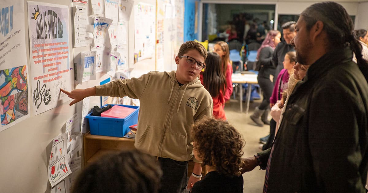 A boy wearing glasses presents a project on a classroom wall to an audience of adults and children during what appears to be a school event, offering exhibition tips that could be especially helpful for new teachers. Other families are visible in the background.