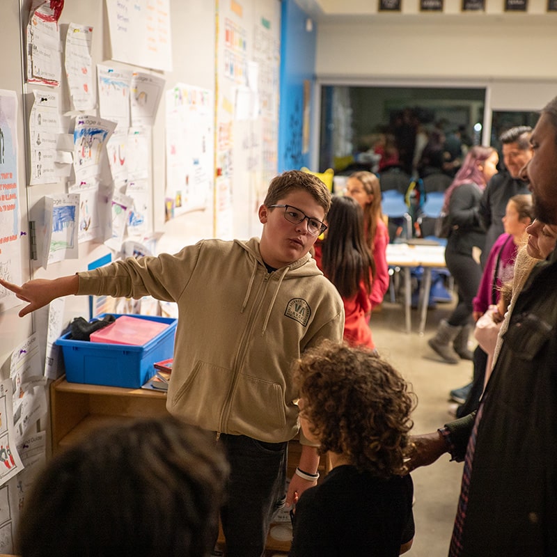 A boy wearing glasses and a beige hoodie stands by a classroom wall covered in student work, pointing to a paper as he shares exhibition advice with new teachers and presents to a group of children and adults during a school event or open house.