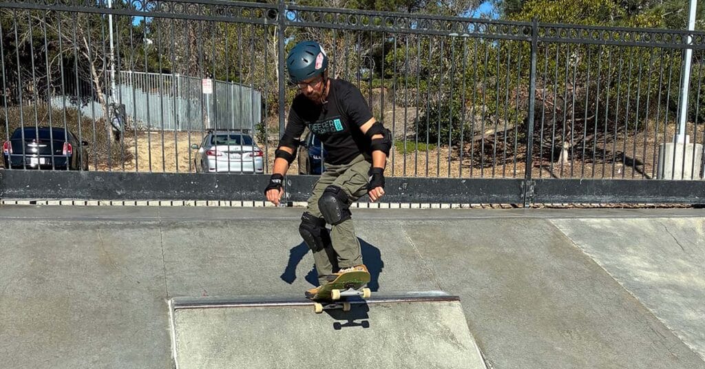 A person wearing a helmet and protective gear is skateboarding at a skatepark, preparing to ride down a ramp. The scene, featured in Unboxed Issue 29, includes a metal fence and parked cars in the background.
