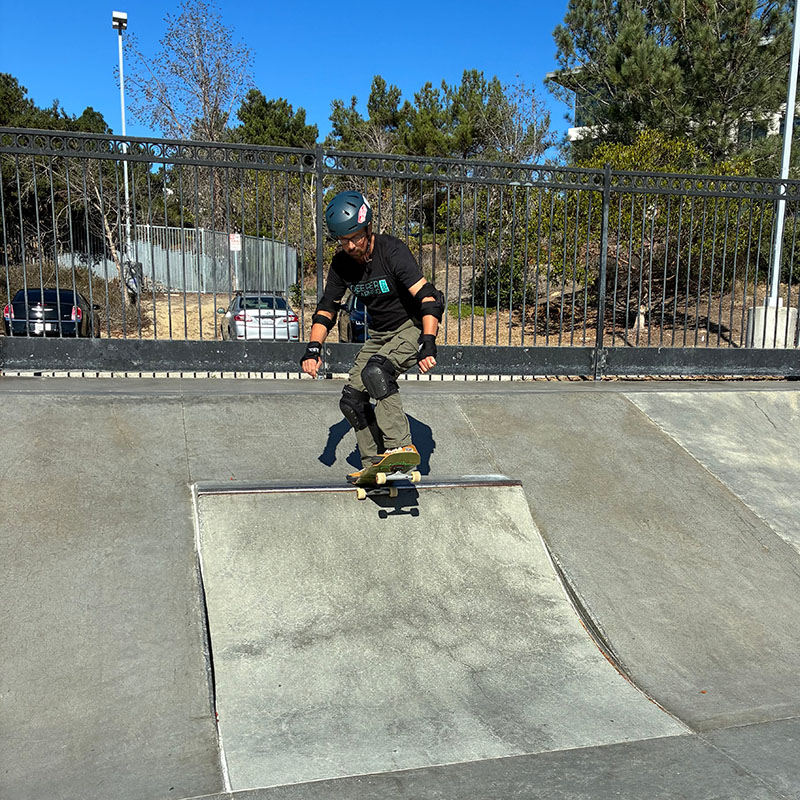 A skateboarder wearing a helmet and pads is skating down a concrete ramp at an outdoor skate park, featured in Unboxed Issue 29, with trees and parked cars in the background.