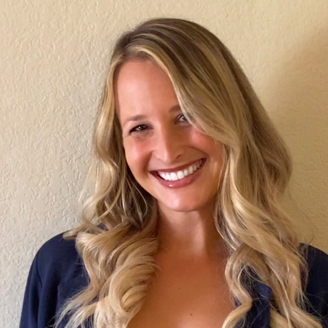 Carly Sumrow Cox, a woman with long, wavy blonde hair, smiles at the camera. She is wearing a dark top and standing in front of a plain, light-colored wall.