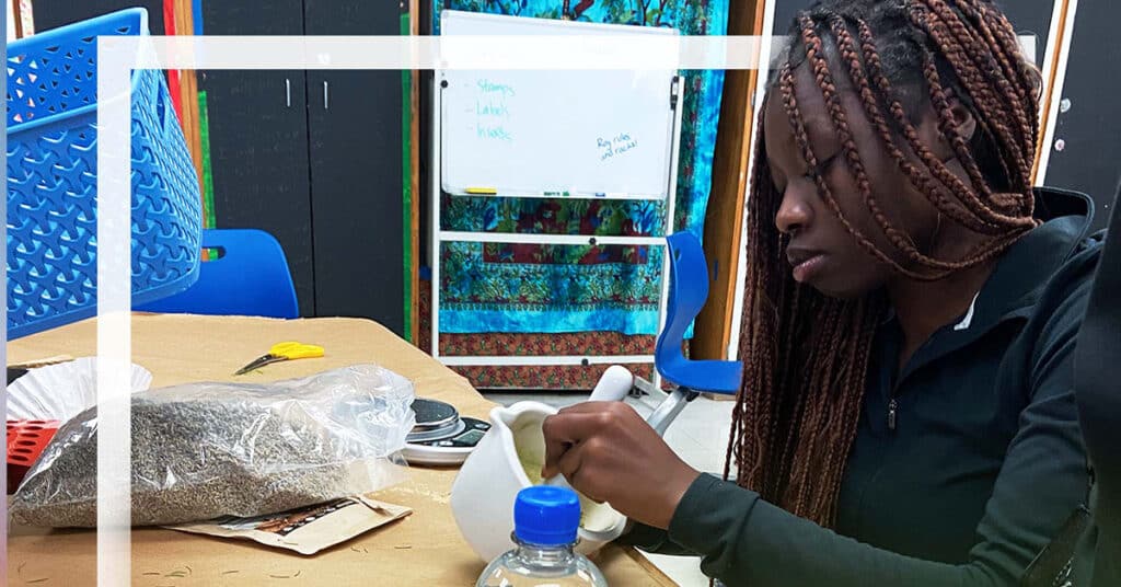 A person with long braids grinds something in a mortar and pestle at a table filled with items used in cosmetics, including a plastic bag of seeds, a coffee filter, scissors, and a bottle. A whiteboard and colorful fabric are in the background.