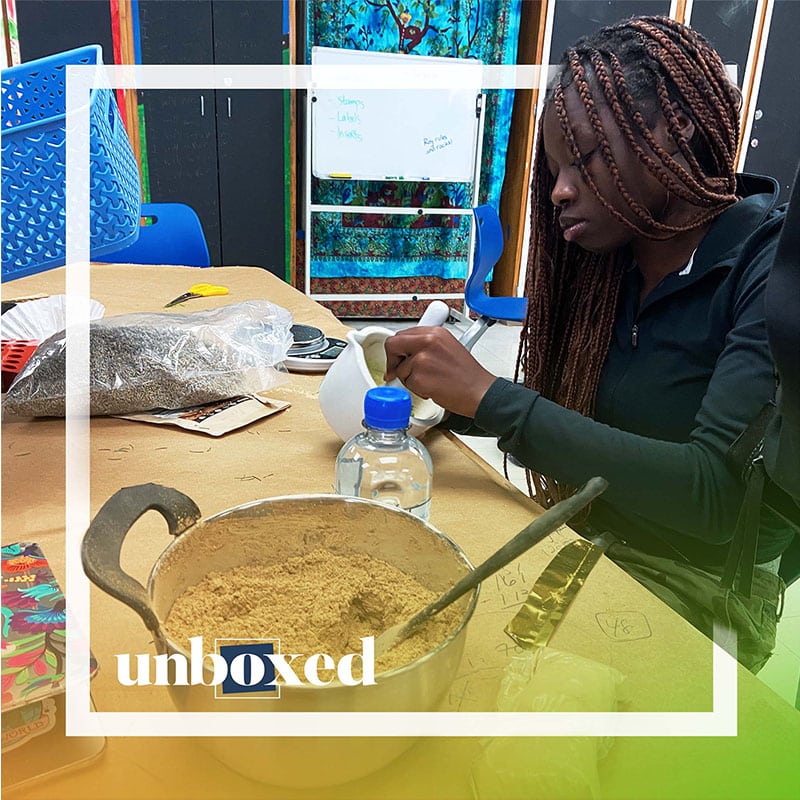 A young woman with long braids mixes ingredients in a white bowl at a table with a pot of brown powder, a water bottle, and craft supplies—showcasing her entrepreneurship in cosmetics. The word unboxed appears in the bottom left corner.