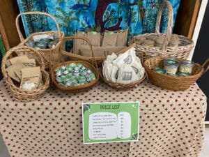 A display of baskets holding candles, jars, tins, and packaged goods is arranged on a star-patterned cloth. A green price list highlights items and prices, showcasing creative entrepreneurship. A colorful backdrop brightens the scene.