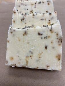 Three bars of handmade soap with visible specks of dried herbs, possibly lavender, arranged in a row on a brown surface. The soap has a creamy color and rustic texture—perfect for those passionate about natural cosmetics.