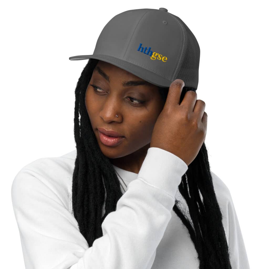 A person with long black hair and a nose piercing wears a gray closed back hat with embroidered hhtgse in blue and yellow, paired with a white sweatshirt, touching the brim of the cap.