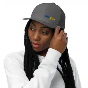 A person with long black hair and a nose piercing wears a gray closed back hat with embroidered hhtgse in blue and yellow, paired with a white sweatshirt, touching the brim of the cap.