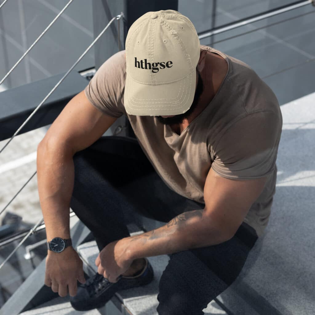 A man sits on outdoor stairs, wearing a distressed fitted cap with the text “hthgse,” a taupe t-shirt, dark pants, and a wristwatch. His head is bowed, and his arms rest on his knees.