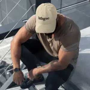 A man sits on outdoor stairs, wearing a distressed fitted cap with the text “hthgse,” a taupe t-shirt, dark pants, and a wristwatch. His head is bowed, and his arms rest on his knees.