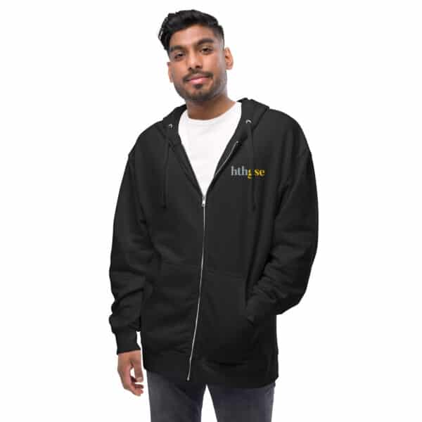 A man wearing a black fleece zip-up hoodie with the word htlqse printed in small, white and yellow letters on the left chest stands with one hand in his pocket against a white background. This unisex hoodie offers both style and comfort.