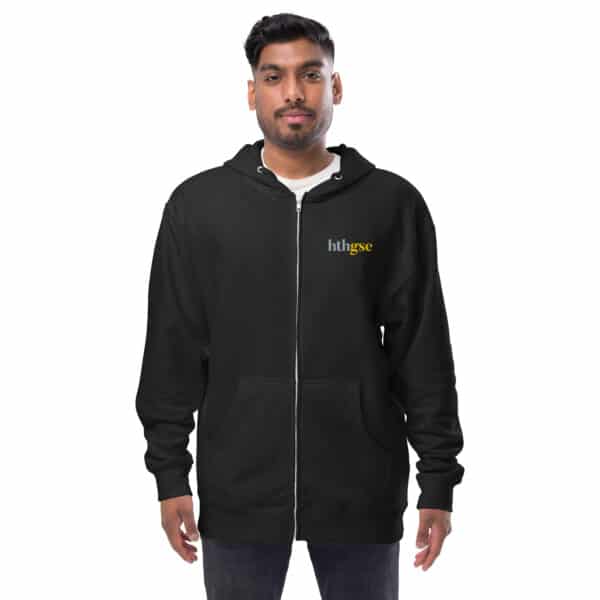 A man with short dark hair and a beard wears a black unisex fleece zip-up hoodie with htb|gse embroidered in small yellow and white letters on the left chest, standing against a plain white background.