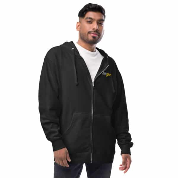 A man stands facing forward, wearing a black unisex zip-up hoodie with a small yellow logo on the chest over a white t-shirt, and dark jeans. He has short dark hair, a trimmed beard, and he is smiling slightly.