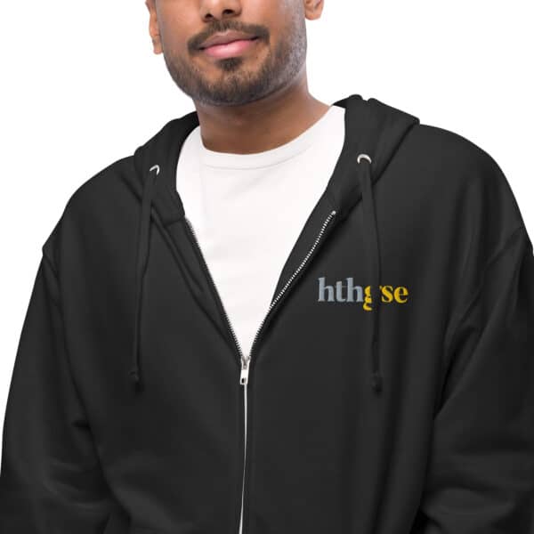 A person wearing a black unisex fleece zip-up hoodie with the word hthgse printed in gray and yellow, over a white t-shirt. Only the lower half of their face is visible.