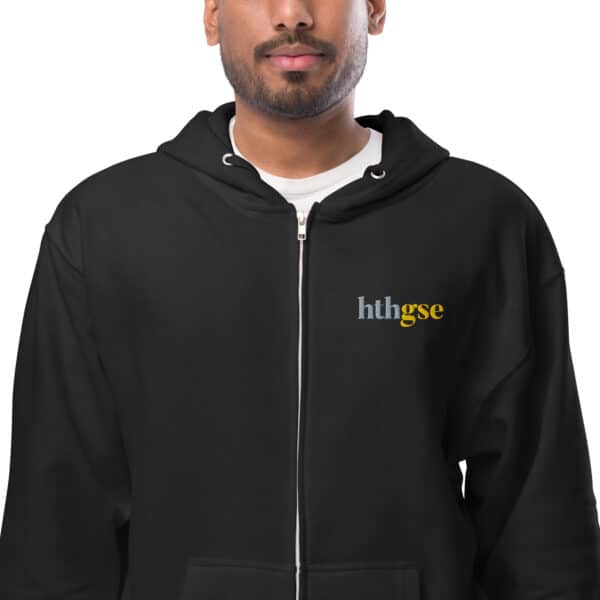 A person wearing a black unisex fleece zip-up hoodie with “hthgse” embroidered on the left chest in white, gray, and yellow. Only the lower face and upper body are visible.