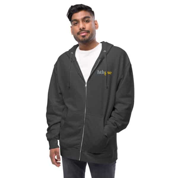 A man wearing a dark gray fleece zip-up hoodie with hthse embroidered on the left chest in white and yellow, over a white t-shirt, stands with one hand in his pocket against a plain white background.