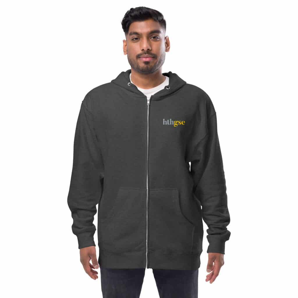 A man stands facing forward, wearing a dark embroidered fleece zip-up hoodie with the word huhsee in small yellow and white letters on the left chest. The background is plain white.