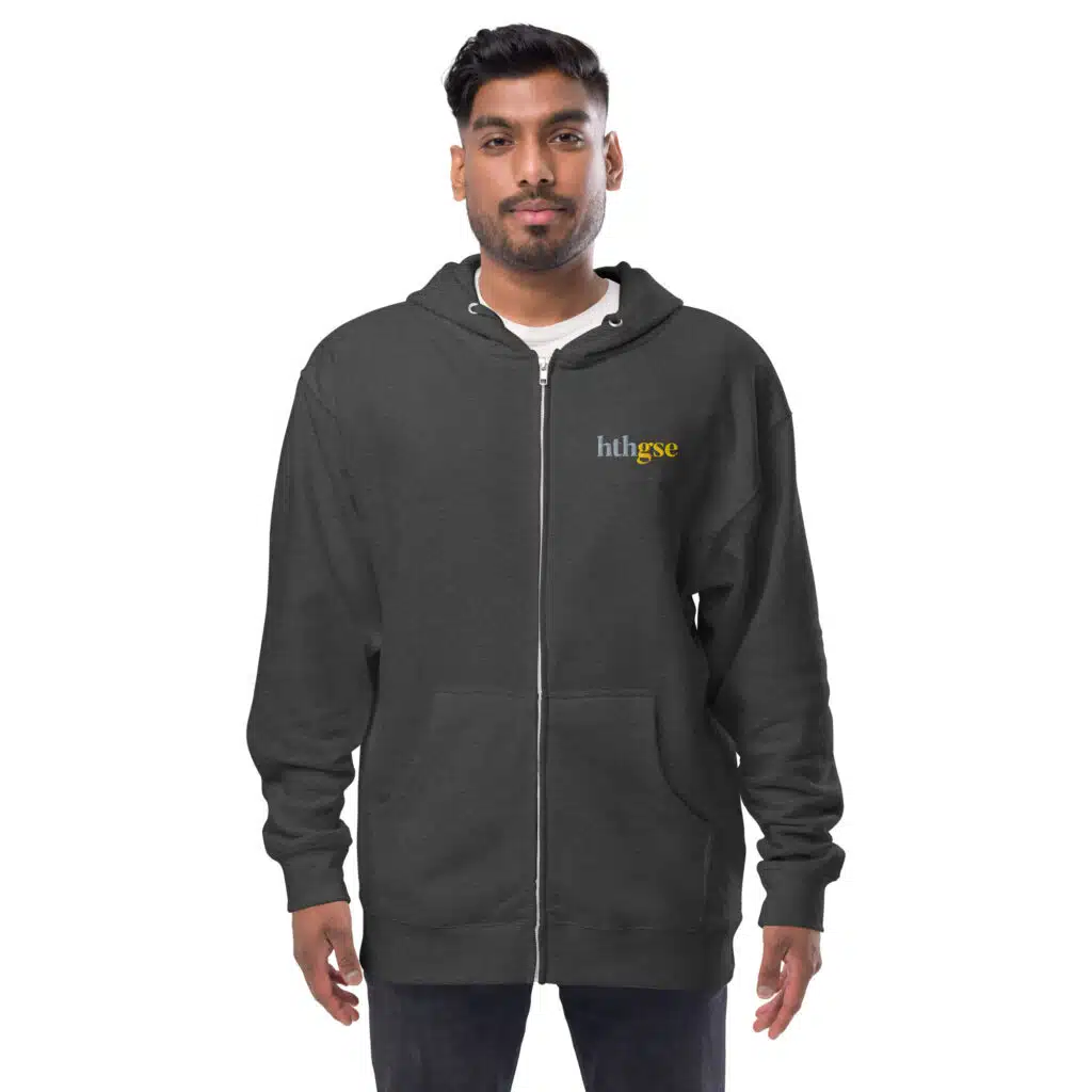 A man stands facing forward, wearing a dark embroidered fleece zip-up hoodie with the word huhsee in small yellow and white letters on the left chest. The background is plain white.