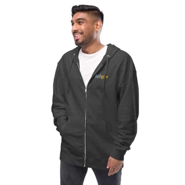 A man stands smiling with his hands in the pockets of a dark gray unisex fleece zip-up hoodie over a white t-shirt. The hoodie features small embroidered text on the left chest. He has short black hair and a trimmed beard.