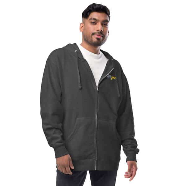A man wearing a dark gray unisex fleece zip-up hoodie with a small embroidered yellow and white logo on the chest stands with a relaxed expression, hands slightly apart, over a white t-shirt and dark pants against a plain white background.