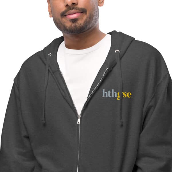 A person wears a dark gray unisex zip-up hoodie over a white shirt. The fleece hoodie features "hthgse" printed on the left chest in blue and yellow. Only the upper body is visible as the person faces forward.