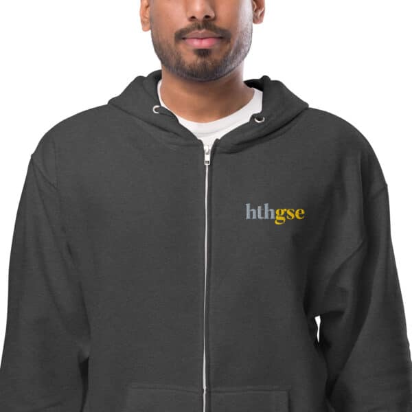 A person wearing a dark gray unisex fleece zip-up hoodie with the letters hthgse printed on the left chest in white and yellow text. The hoodie is worn over a white t-shirt.
