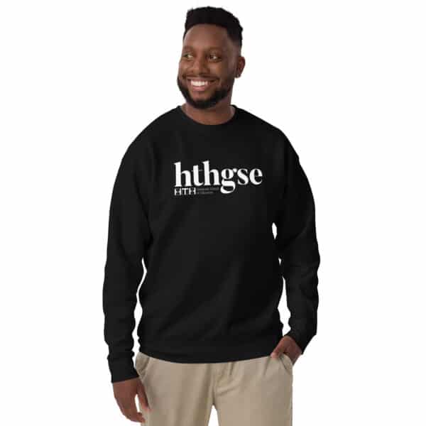 A smiling man wearing a black crew neck sweatshirt with a large white logo and smaller text beneath it, standing with one hand in his pocket against a plain white background.