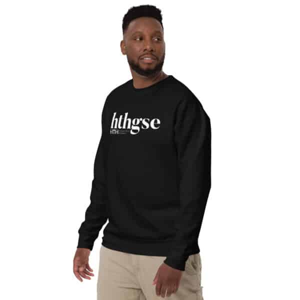 A man wearing a Unisex Crew Neck Sweatshirt with the text hthgse in white and light-colored pants stands against a plain white background, looking slightly to his left with a relaxed expression.