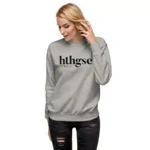A person with long blonde hair wearing a unisex crew neck gray sweatshirt that reads hthgse in large black letters, standing and looking down with one hand touching their hair, dressed in ripped black jeans.