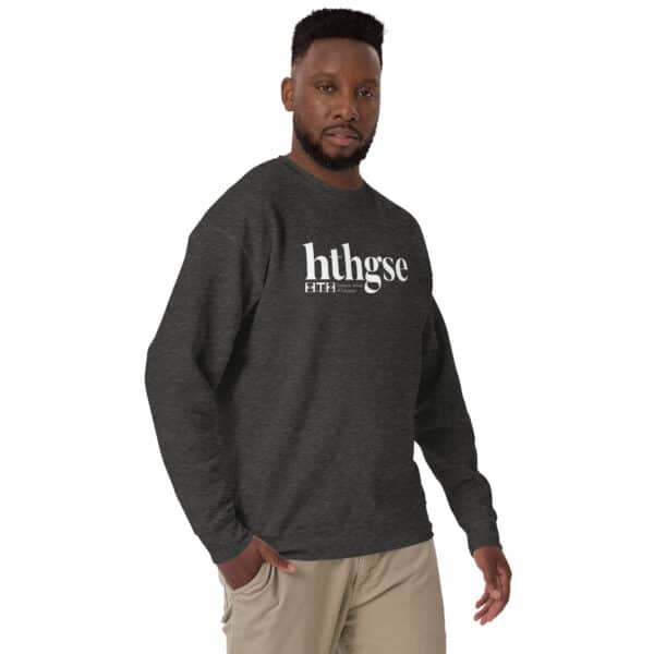 A man wearing a dark gray Unisex Crew Neck Sweatshirt featuring a white logo and text reading hthgse and F I T H stands against a plain background, looking at the camera with one hand in his pocket.