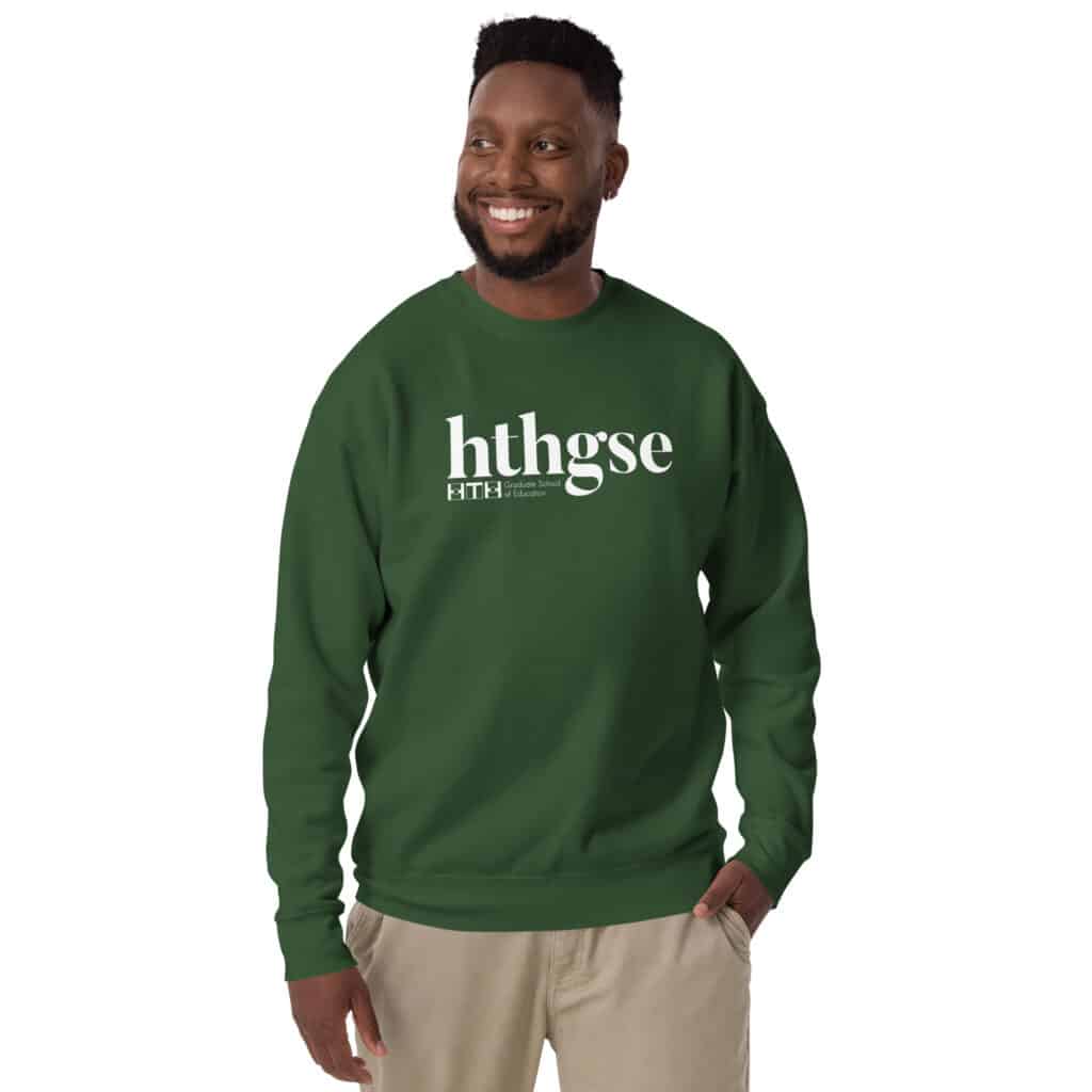 A smiling man with short hair and a beard wears a unisex crew neck sweatshirt, featuring “hthgse” in large white letters on the front. He stands against a plain white background with one hand in his pocket.